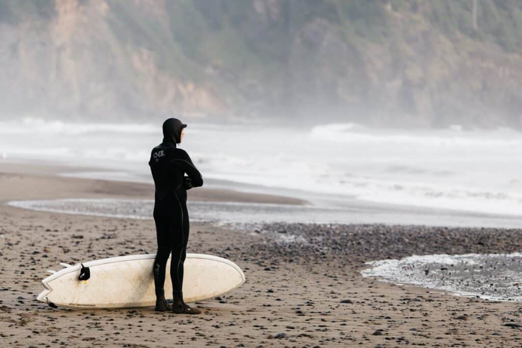 A person in a wet suit standing on the beach ,looking at the water, with a surf board next to them