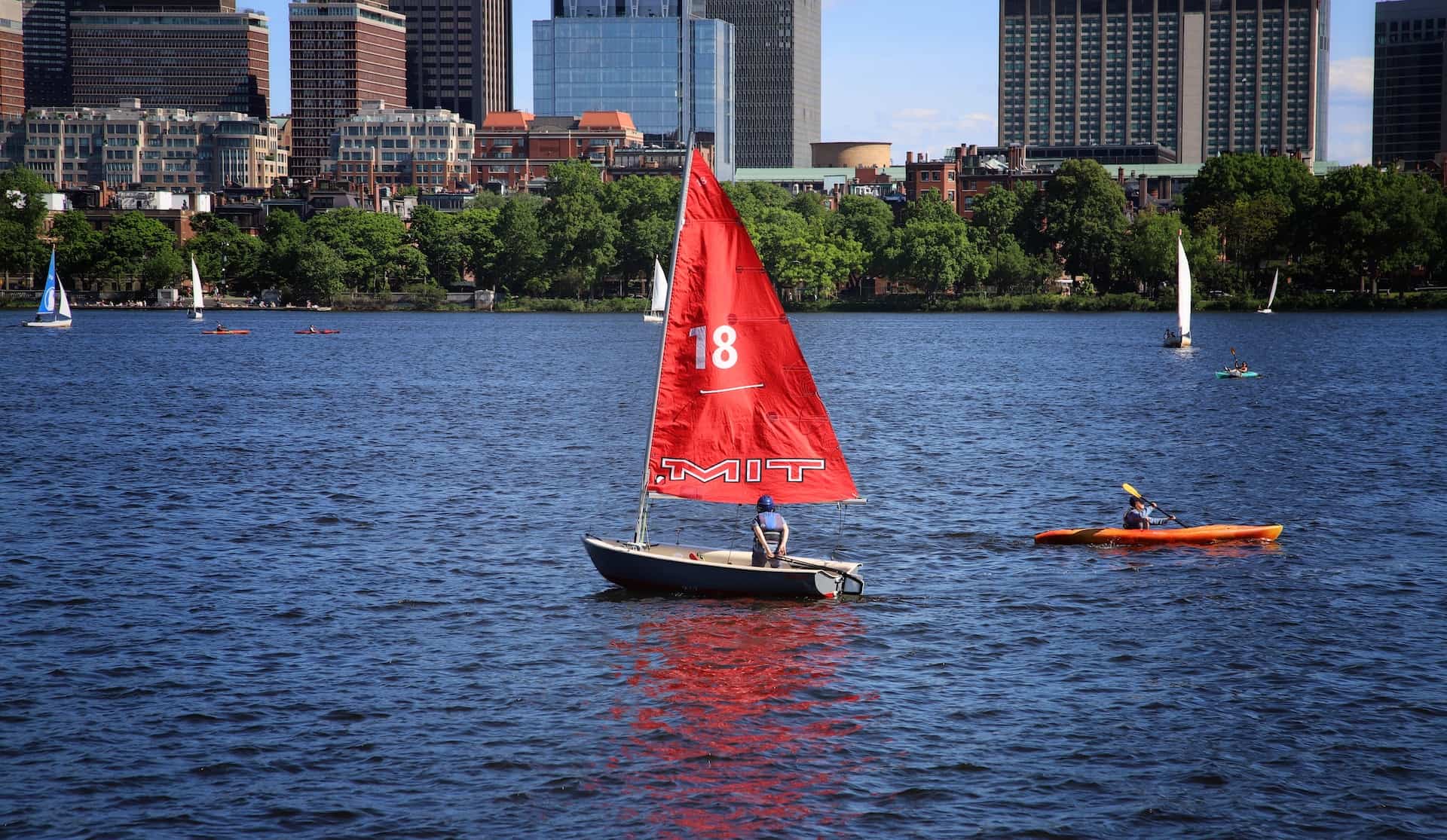 Image of a Sailing Dinghy on a still river with a red sail and a Kayak going past it